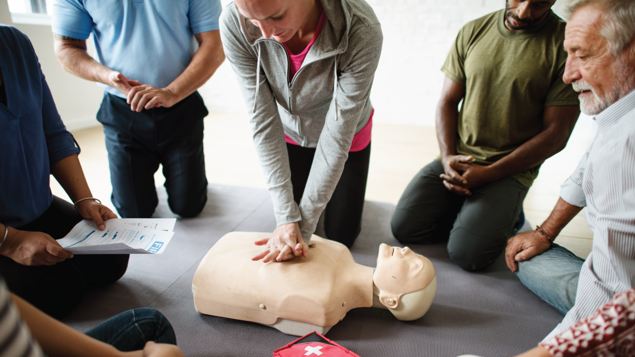 A diverse group of people kneels around a CPR manikin. A woman in the center performs chest compressions. A man holds a CPR instruction sheet. A red medical kit with a white cross is on the floor nearby.
