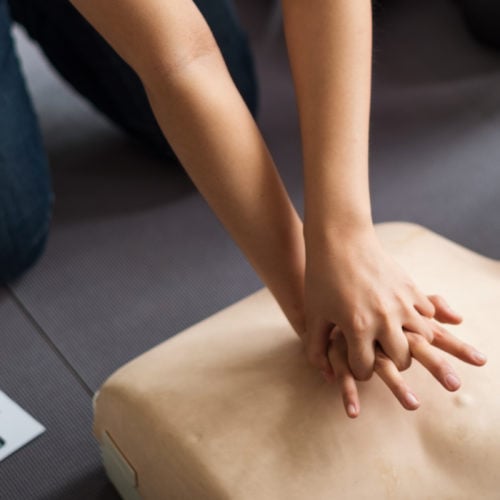 Person practicing CPR on a mannequin, pressing down on the chest with interlocked hands. A first aid manual is visible on the side, and the setting appears to be an indoor training session.
