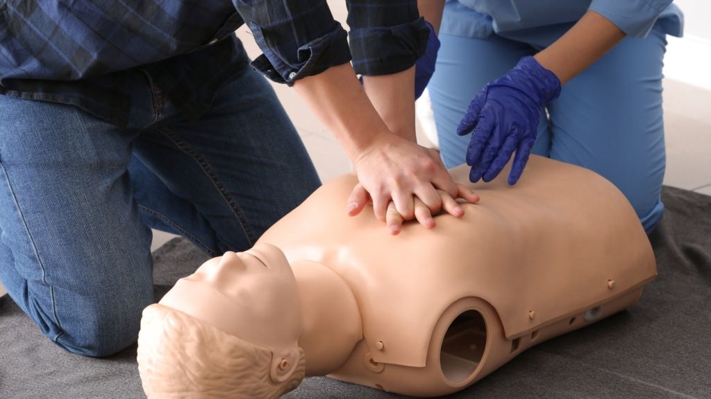 A person wearing blue gloves performs CPR on a medical training mannequin. Another person kneels beside them, observing or assisting. The mannequin is lying on a gray surface.