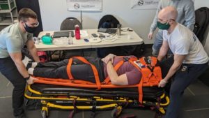 Three people are securing a person on a stretcher with orange straps. The person on the stretcher is lying down with a neck brace. A table with medical equipment is in the background. All individuals are wearing masks.