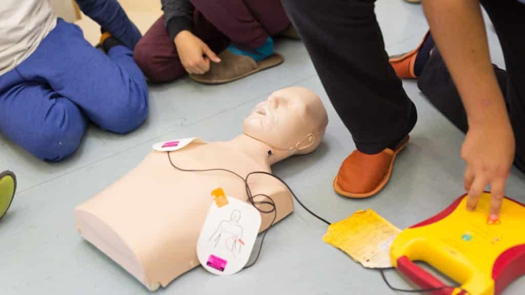 A group of people is practicing CPR on a training mannequin. The mannequin has defibrillator pads attached to its chest, and a defibrillator device is placed nearby. The focus is on learning emergency response skills.