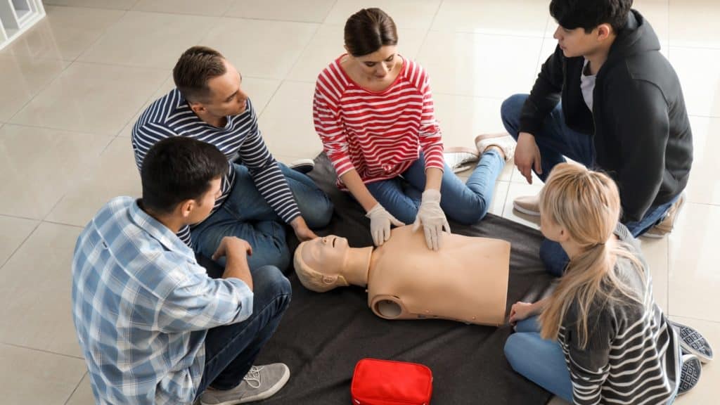Five people are seated on the floor around a CPR mannequin. One person is demonstrating chest compressions, wearing gloves. A red first aid kit is nearby. The group is in a well-lit indoor space with a tiled floor.