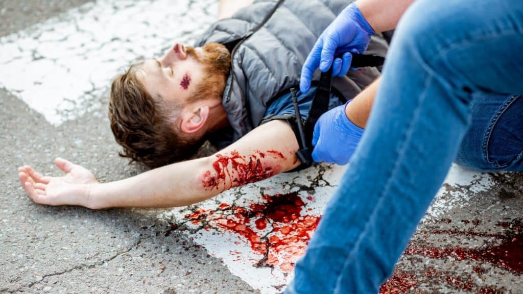 A person with a bloody arm injury lies on the ground. Another individual wearing blue gloves applies a tourniquet to the injured arm. There is a pool of blood on the pavement nearby.