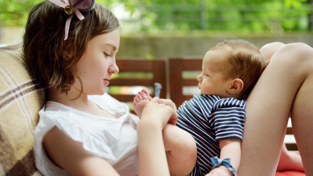 A young girl with a pink bow in her hair sits on an outdoor patio, lovingly holding and looking at a baby in a striped onesie. The baby lies on her bent legs, and both appear content in a serene setting.