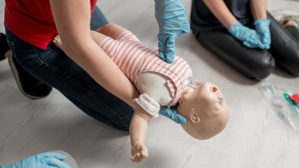 A person in a red shirt and blue gloves is demonstrating CPR on a baby mannequin wearing a striped outfit. Another person, also wearing blue gloves, sits nearby on the floor. Medical supplies are visible on the floor.