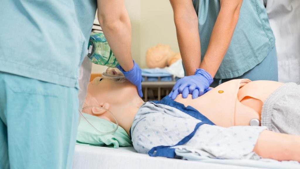 Medical training scenario with a mannequin. A person in blue gloves performs chest compressions while another holds an oxygen mask over the mannequin's face. Both are wearing scrubs, simulating an emergency medical procedure.