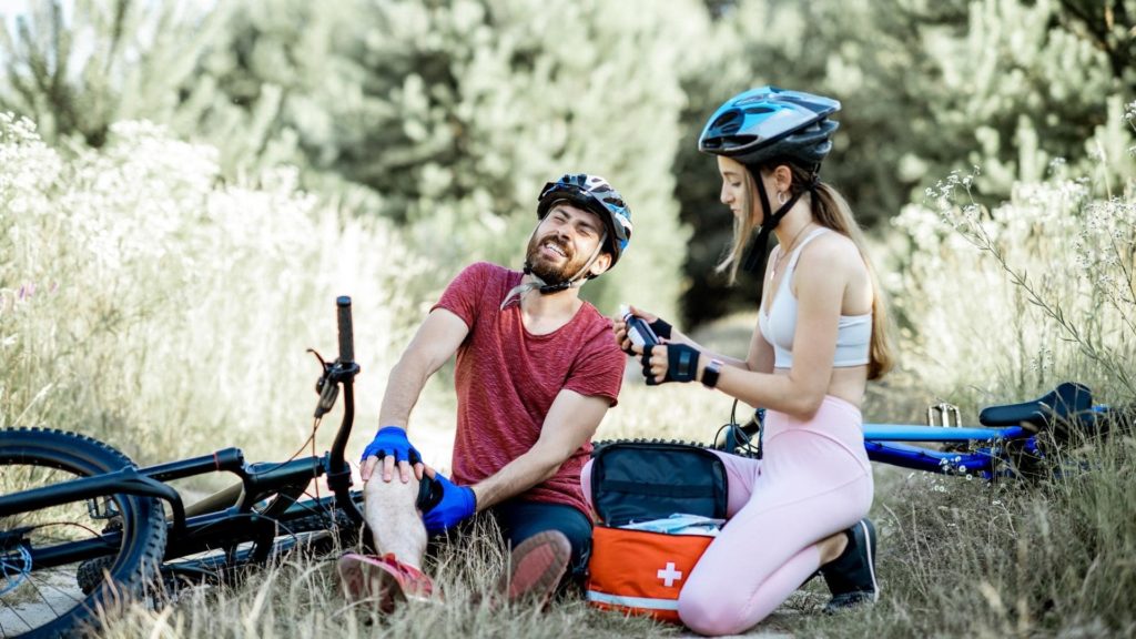A man sits on the ground next to a fallen bike, holding his knee with a pained expression. A woman in athletic gear tends to him with a first aid kit. Both wear helmets, and they're surrounded by lush greenery.