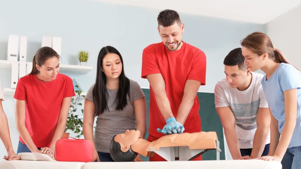 A group of five people watches as a man in a red outfit demonstrates CPR on a mannequin. The scene takes place indoors, with a red first aid kit visible on a table nearby.