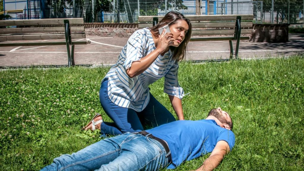 A woman in a striped shirt is kneeling on grass, talking urgently on her phone. A man in a blue shirt and jeans is lying on the grass, appearing unconscious. It's a sunny day, with benches and a fenced area in the background.