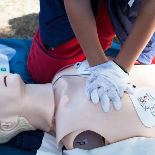 A person is performing CPR on a mannequin outdoors. They are applying chest compressions, wearing gloves. Nearby, a defibrillator machine is visible on the ground. The scene suggests a first aid training session.