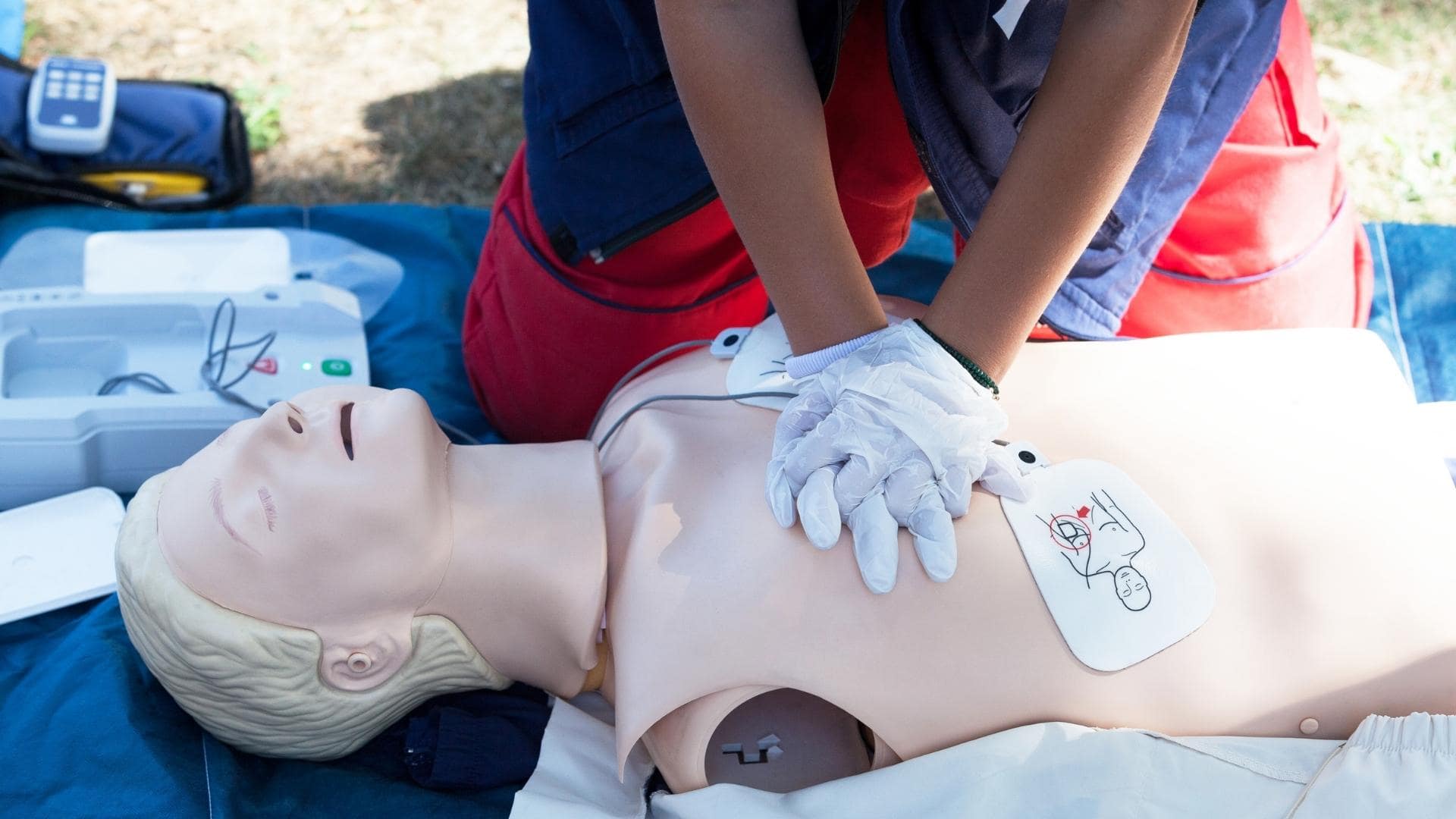 A person is performing CPR on a mannequin outdoors. They are applying chest compressions, wearing gloves. Nearby, a defibrillator machine is visible on the ground. The scene suggests a first aid training session.