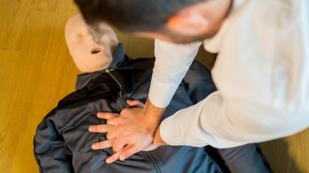 A person is practicing CPR on a dummy. The dummy is dressed in a dark jacket and lying on a wooden floor. The individual performing CPR is pressing down on the dummy's chest, focusing on the technique.