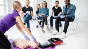 A group of six people sits on chairs, attentively watching an instructor demonstrate CPR on a mannequin. They are wearing gloves, and an automated external defibrillator is placed nearby on the floor.