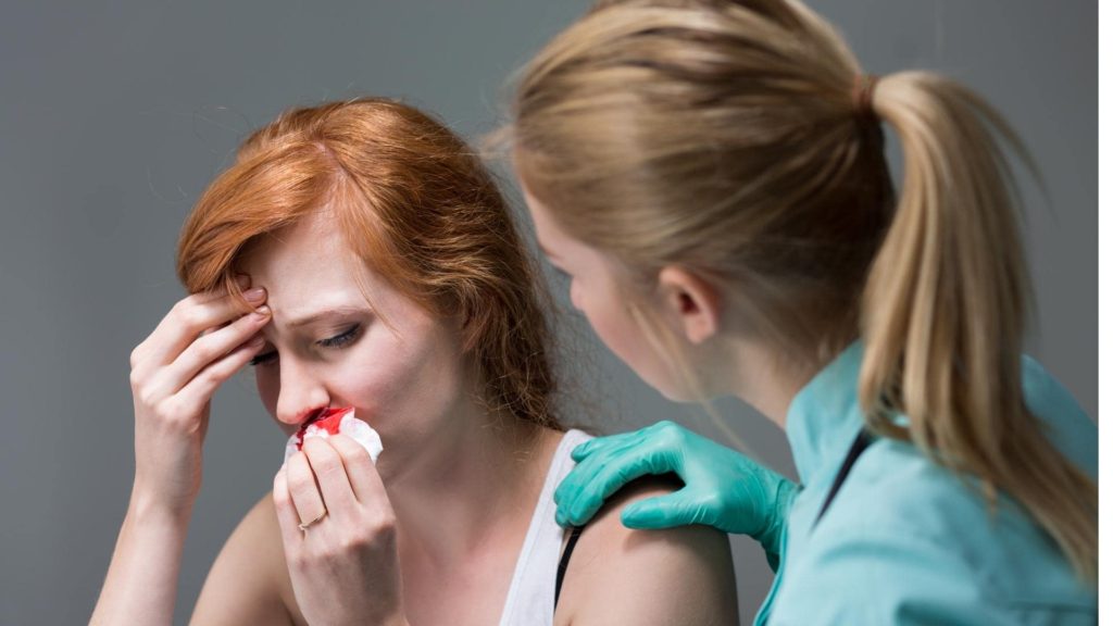 A woman with red hair holds a tissue to her bleeding nose, showing discomfort. Another woman with blonde hair and a ponytail, wearing green gloves, gently places a hand on her shoulder, providing support and assistance.