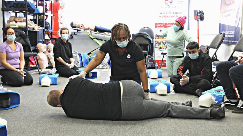 A group of people attending a first aid training session. A trainer demonstrates a recovery position on a person lying on the floor. Participants watch attentively, surrounded by CPR mannequins and medical equipment. Everyone is wearing masks.