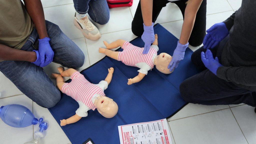 People are kneeling on the floor, wearing blue gloves, practicing CPR on two baby mannequins dressed in striped outfits. A set of instructions and a medical bag are nearby.