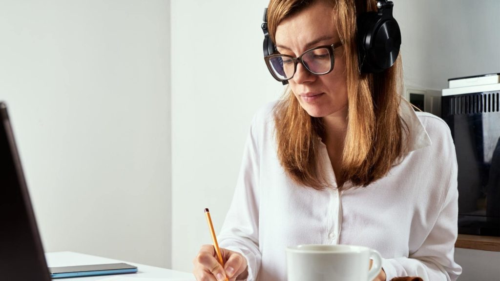 Woman with long hair and glasses wearing headphones sits at a desk, writing with a pencil on paper. An open laptop is in front of her, and a large mug is on the table. She appears focused in a bright indoor setting.