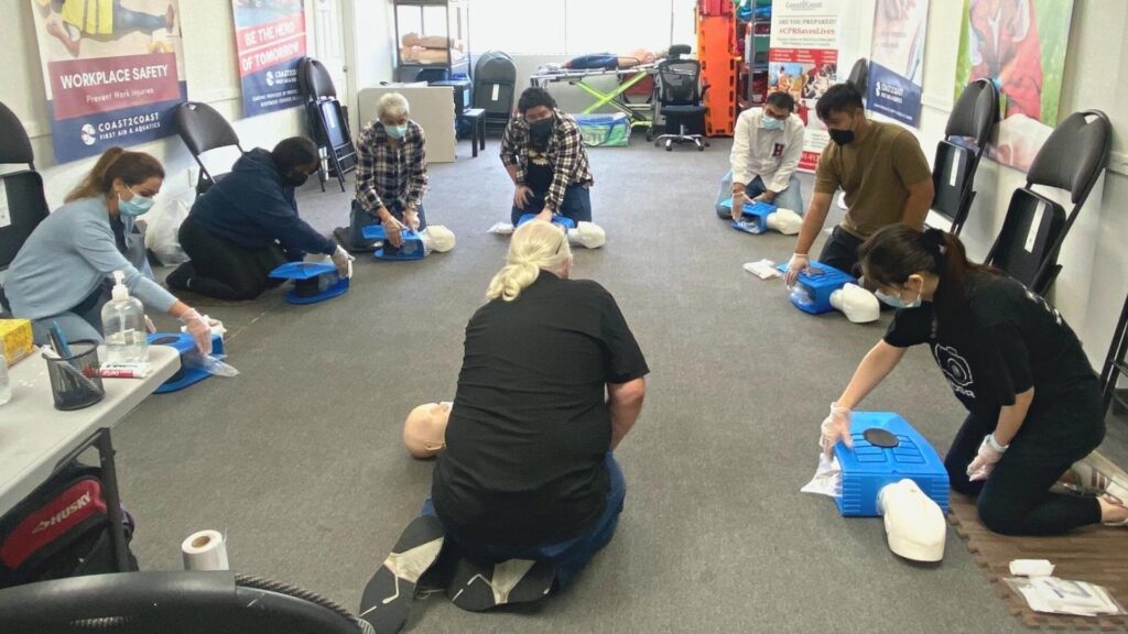 People are practicing CPR on training manikins in a classroom setting. They are wearing masks and gloves, arranged in a circle, with a safety poster on the wall. Tables with equipment are visible in the background.