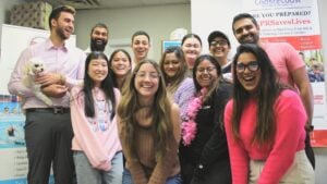 A group of thirteen people, diverse in appearance, smiling and posing indoors. One person on the left holds a small white dog. Behind them are posters about training and safety.