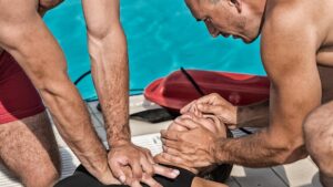 Two lifeguards perform CPR on an unconscious person by a pool. One lifeguard applies chest compressions while the other holds the person's head, preparing to give rescue breaths. A rescue tube is visible in the background.