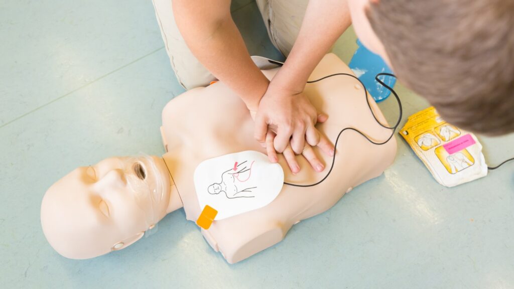 The image shows a person practicing CPR on a training mannequin. They are performing chest compressions while an automated external defibrillator (AED) is attached to the mannequin.