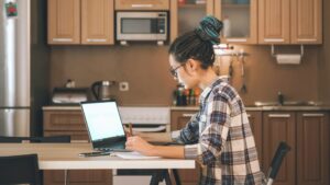Woman working on a laptop at a kitchen table, representing a student taking the online portion of a blended first aid course in Brampton