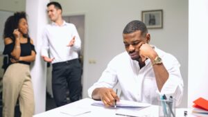Office worker looking stressed at his desk while colleagues talk in the background, representing mental health and wellness in the workplace challenges