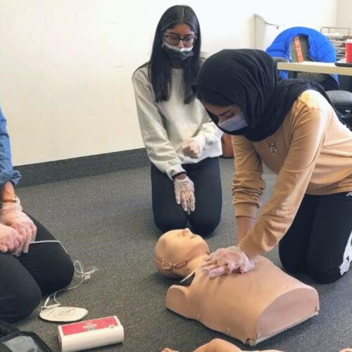 Four individuals are participating in a CPR training session. One person is practicing chest compressions on a CPR manikin, while others observe. An AED device is visible on the floor next to them.