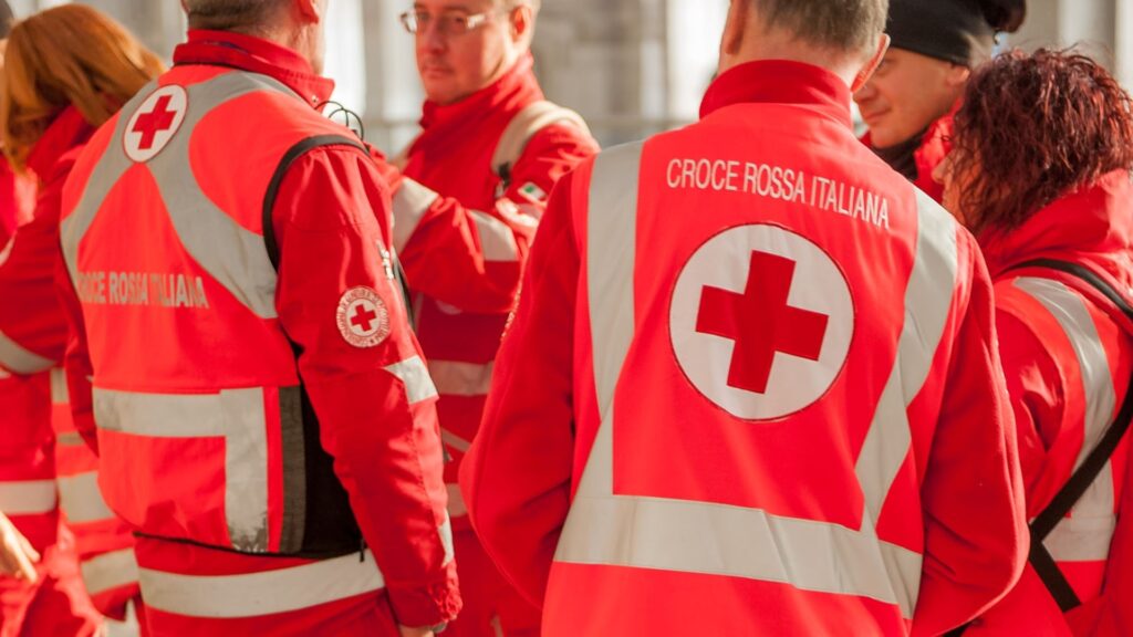 A group of people wearing red uniforms with reflective vests and the "Croce Rossa Italiana" logo, featuring a red cross, on their backs. They seem to be in discussion in an outdoor setting.