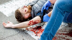 A person with a bloody arm injury lies on the ground. Another individual wearing blue gloves applies a tourniquet to the injured arm. There is a pool of blood on the pavement nearby.