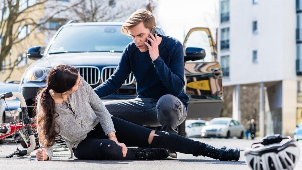 A man is kneeling on the road beside a woman who appears to have fallen off her bicycle. He is on the phone, looking concerned. A car with an open door is in the background, along with another bicycle and buildings.