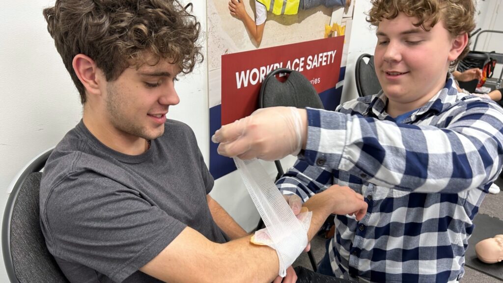 Two young men, one with curly hair in a gray shirt sitting, the other in a checkered shirt standing, practicing bandaging an arm. The standing person is wearing gloves, and there is a "Workplace Safety" poster in the background.