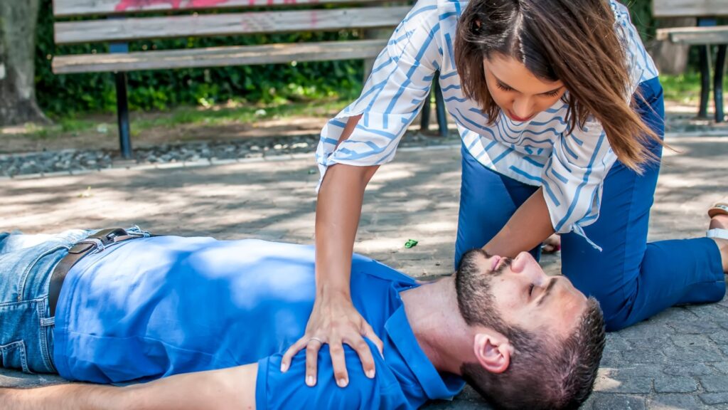 A woman kneels on the ground, performing CPR on an unconscious man lying on his back in a park setting. She is applying chest compressions. Both individuals are dressed casually. A wooden bench and trees are in the background.