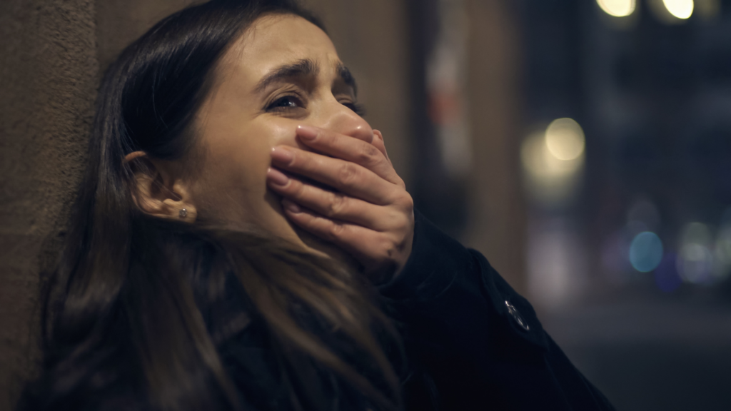A woman with long hair stands against a dimly lit wall, covering her mouth with her hand. Her eyes are wide open, and she appears to be in distress. The background is blurry, suggesting a nighttime urban setting.