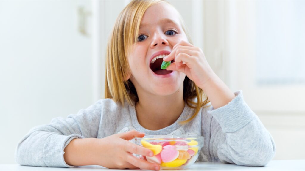 A child with long blonde hair is seated at a table, wearing a gray sweater. They are smiling and holding a colorful gummy candy near their mouth. A glass bowl filled with various candies is in front of them.