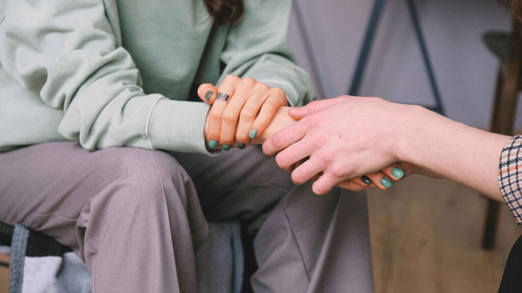 Two people sitting close together, one wearing a green sweater and gray pants, holding hands with another person. Both have turquoise-painted nails. The scene conveys a sense of comfort and support.