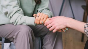 Two people sitting close together, one wearing a green sweater and gray pants, holding hands with another person. Both have turquoise-painted nails. The scene conveys a sense of comfort and support.