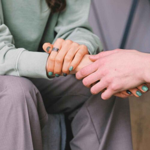 Two people sitting close together, one wearing a green sweater and gray pants, holding hands with another person. Both have turquoise-painted nails. The scene conveys a sense of comfort and support.