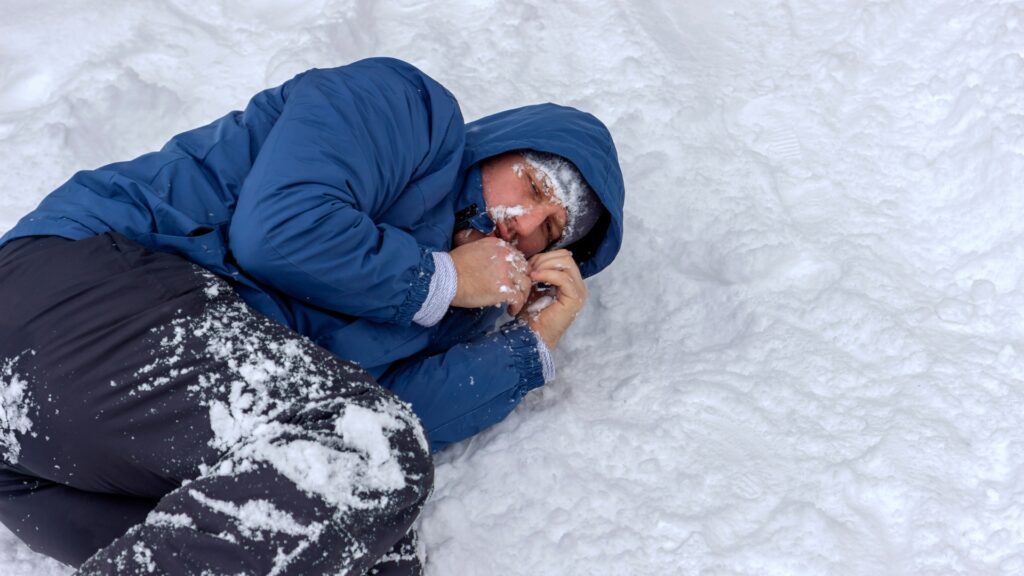 A person wearing a blue jacket and black pants is lying on snow-covered ground. Their face is covered in snow and they are holding a blue object near their mouth. The environment suggests cold, wintry conditions.