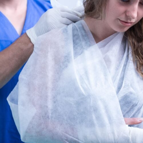 A person in a blue medical uniform is assisting a woman with her arm in a cast and sling. The woman, with curly hair, looks down. They are indoors, in what appears to be a medical setting.
