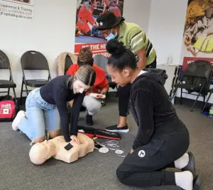 A student practicing chest compressions on an adult manikin while the instructor observes, with an AED trainer and first aid kit on the floor