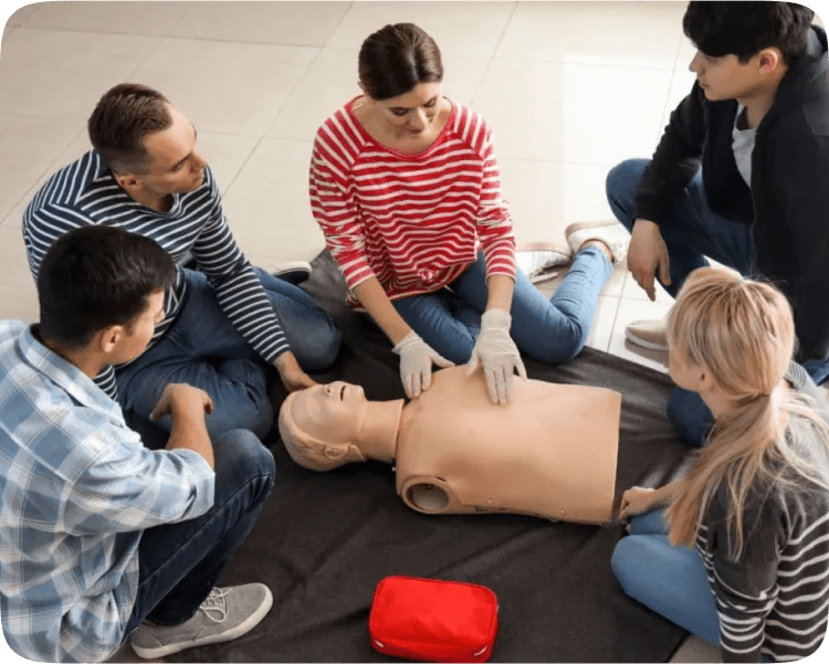 A group of five people is sitting on the floor practicing CPR on a mannequin. One person wearing gloves is demonstrating chest compressions. A red first aid kit is nearby.