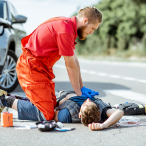 A man in a red jumpsuit performs CPR on another man lying on the road. An open first aid kit is nearby, and a vehicle is parked in the background. The scene appears to be an emergency situation.