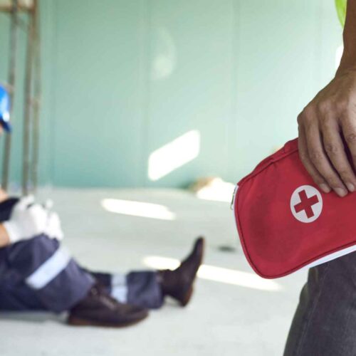 A construction worker sitting on the ground in the background with an injured leg, while another person approaches holding a red first aid kit