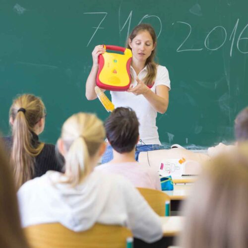 A teacher stands in front of a classroom holding a defibrillator and addressing students. The students, seated and facing the teacher, listen attentively. A chalkboard with writing is behind the teacher in the classroom setting.