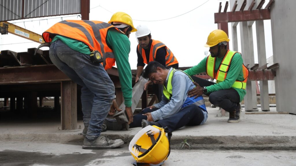 A group of construction workers in safety vests and helmets attending to an injured colleague on a construction site, with a 'SAFETY FIRST' banner in the background 