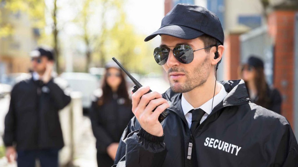 male security guard with hat and glasses talking into a radio with other security guards in the background blurred out