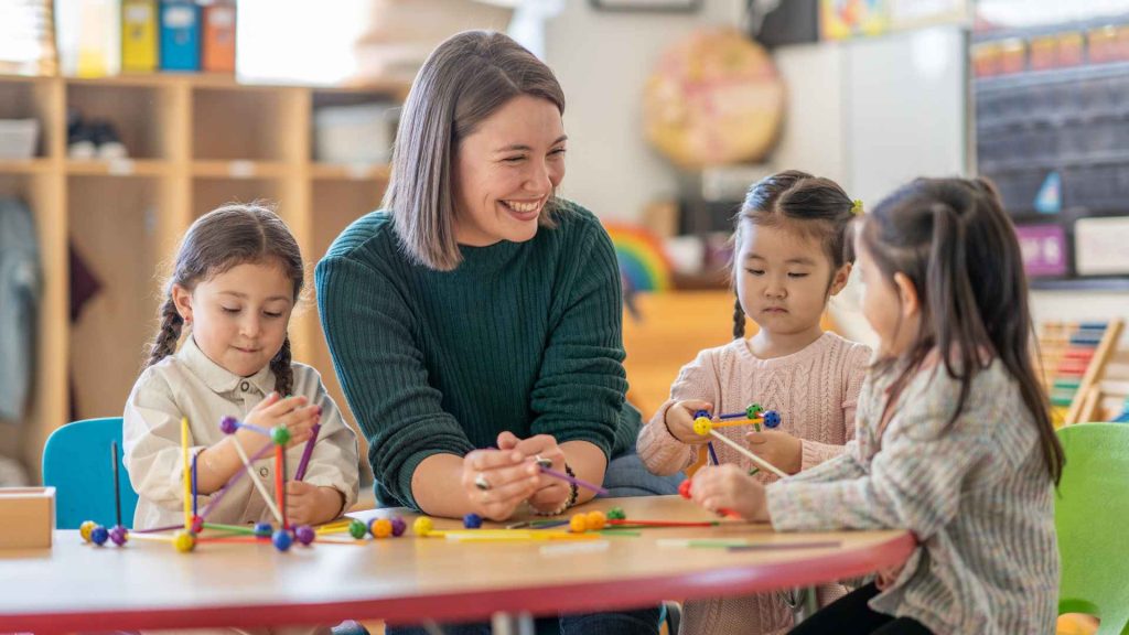female early childhood educator sitting at table with 3 young girls in a classroom