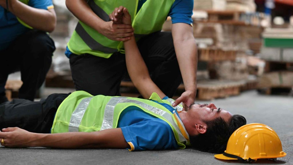 worker in bright yellow vest laying on ground unconscious while another worker checks on him