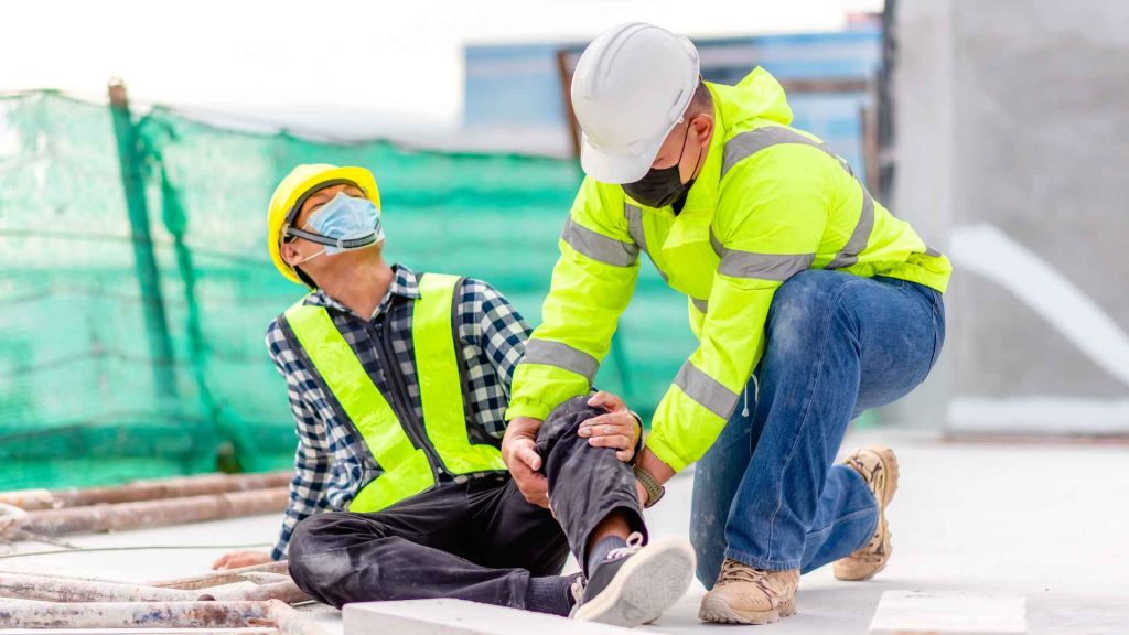 a construction worker sitting on the ground holding his knee in pain with another construction worker holding the knee and looking at the injury